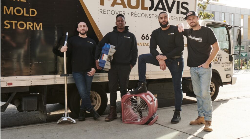 Four Paul Davis restoration team members standing outdoors beside a branded service truck, holding equipment including a squeegee and a drying fan, with services listed as fire, mold, and storm visible on the vehicle.